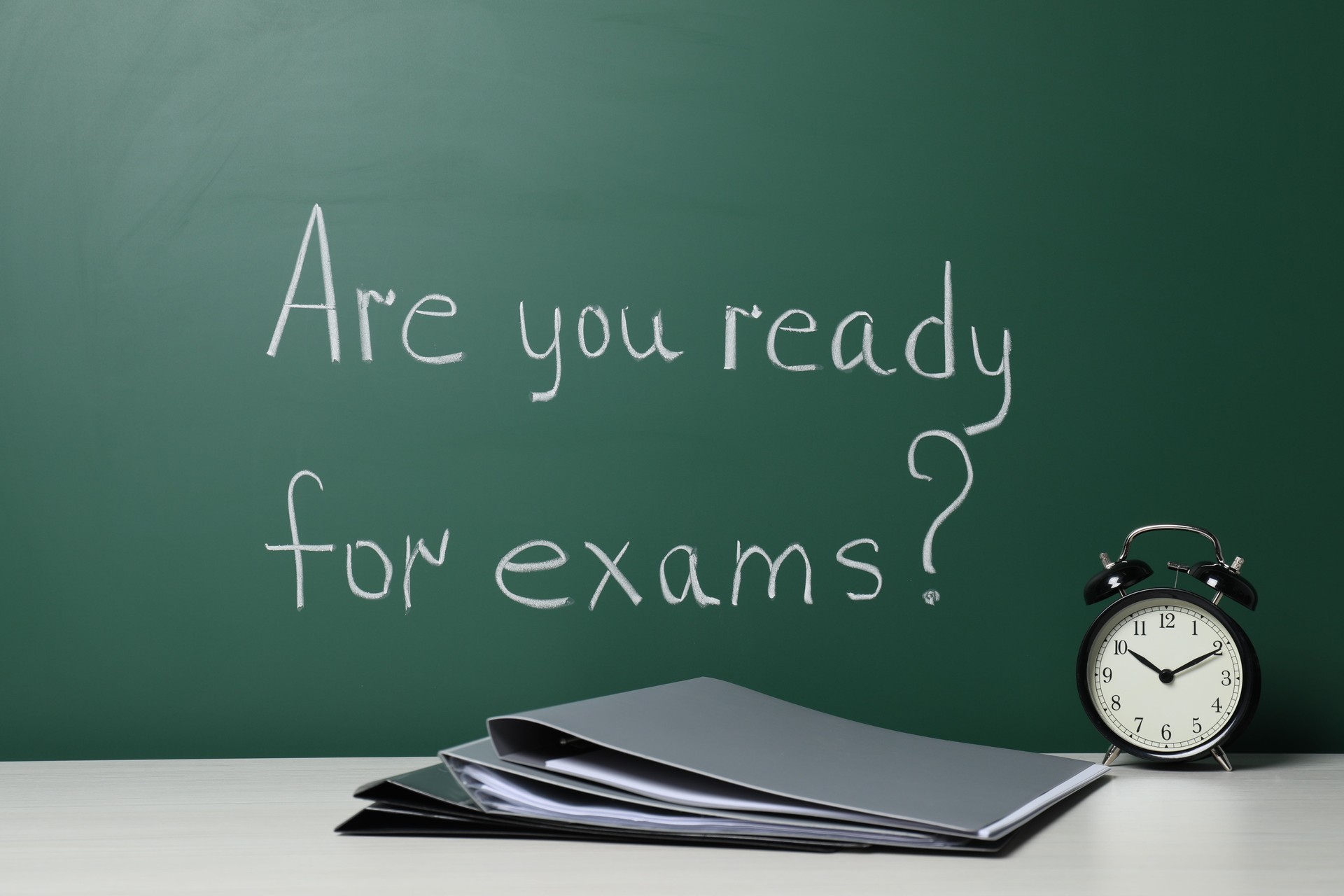 Office folders and alarm clock on white table near chalkboard with phrase Are You Ready For Exams